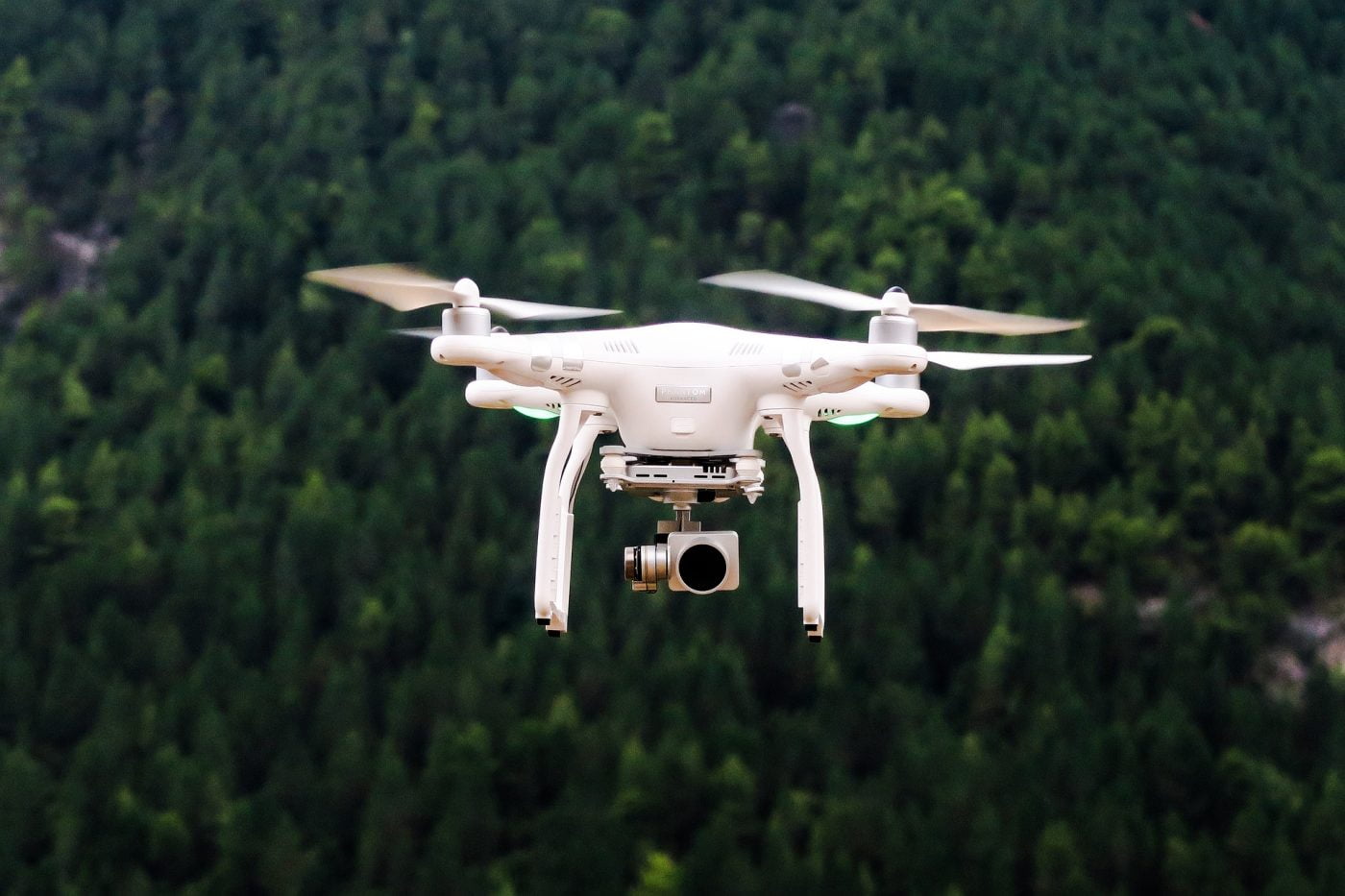 Weiße Drohne in der Luft, im Hintergrund ist ein Wald mit viele Bäumen zu erkennen. Die Drohne hat noch eine kleine Kamera.