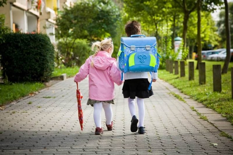 Zwei Kinder mit Rucksack auf dem Rücken. Laufen auf einem Weg an der Straße entlang. Rundum alles grün.