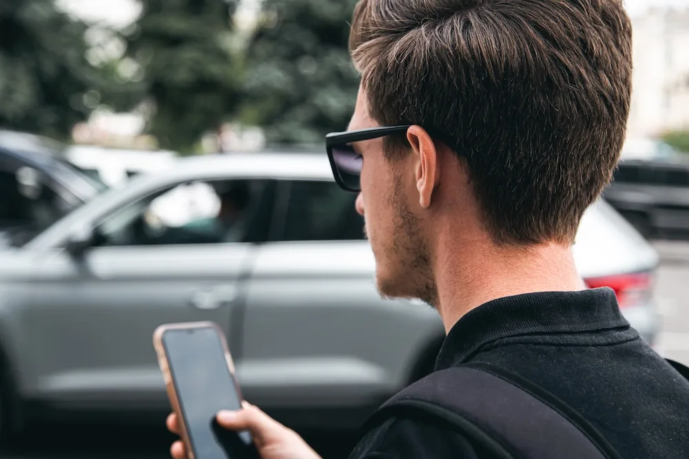 A young man in sunglasses uses a smartphone, close-up, blurred background.