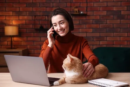 Eine Frau am Telefon schaut auf einen Laptop, neben ihr eine Katze