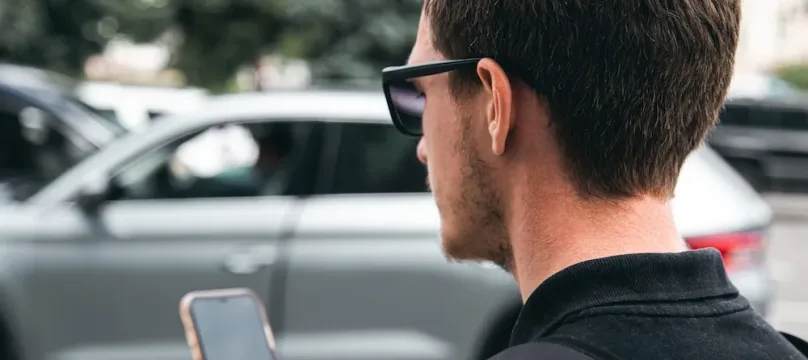 A young man in sunglasses uses a smartphone, close-up, blurred background.